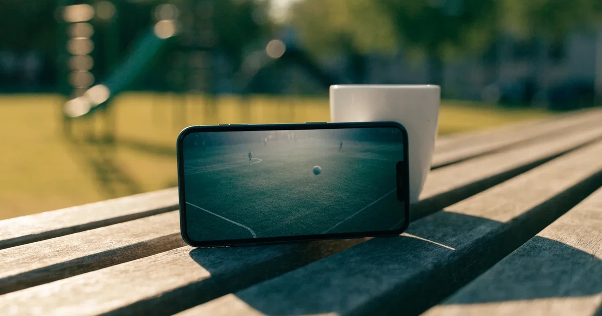 Ein Smartphone mit dunklem Display liegt auf einer Parkbank, daneben ein Kaffeebecher, im Hintergrund ein Spielplatz.