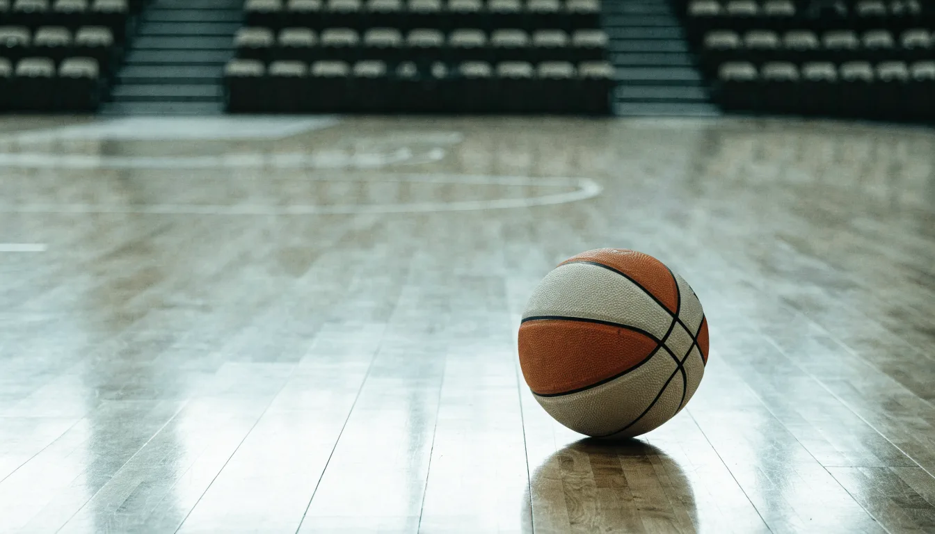 Ein Basketball liegt auf dem Parkett einer leeren Halle, von den Deckenlampen fällt gleichmässiges Licht auf den Boden.