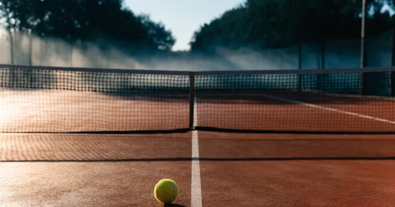 Ein Tennisplatz aus Sand bei Sonnenaufgang mit gespanntem Netz, Nebel zwischen den Bäumen am Rand