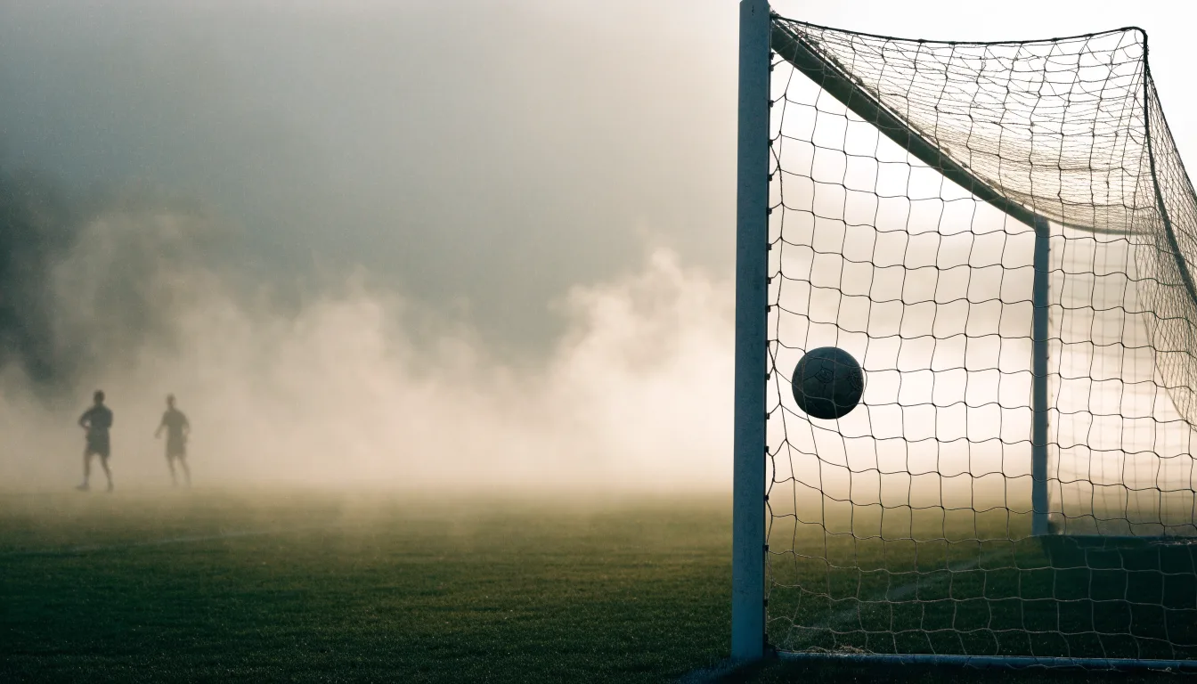 Ein Fussball liegt im Netz hinter einem Tor, weicher Morgennebel hängt über dem Rasen